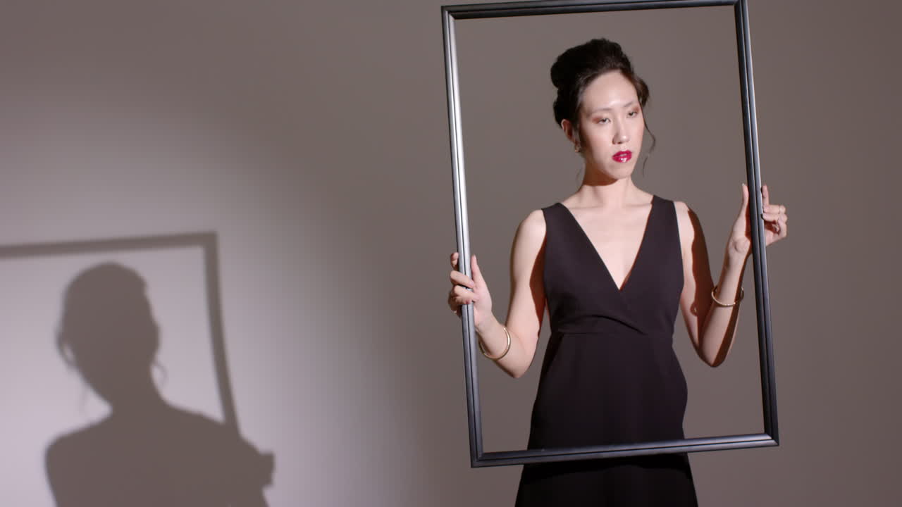 Spotlight casting shadow guiding Asian woman sliding large frame diagonally in studio, copy space