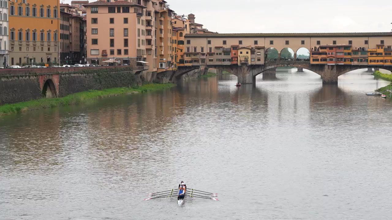 Canottieri in gara scivolano sull'Arno a Firenze, Italia, con il Ponte Vecchio che si erge sullo sfondo. La luce fioca del giorno crea morbidi riflessi e mette in risalto l'iconica architettura rinascimentale