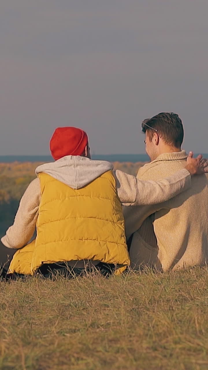 guy in vest sits down on river bank grass edge and hugs frustrated boyfriend in early morning slow motion backside view