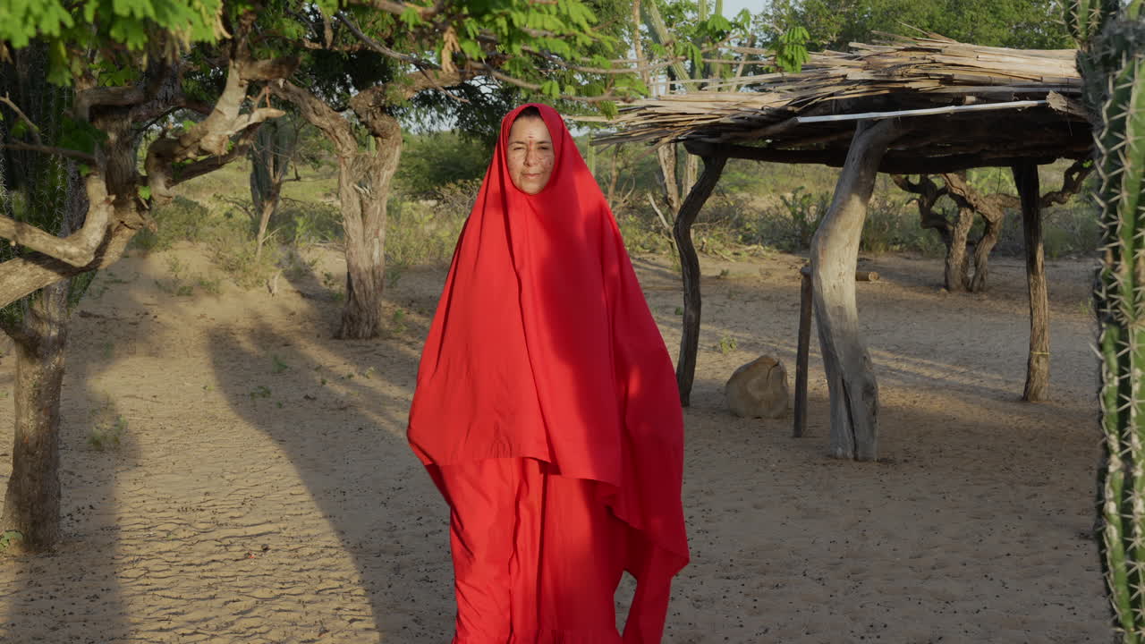 Una donna Wayuu vestita con un vivace mantello rosso cammina attraverso il paesaggio arido e soleggiato de La Guajira, Colombia, con rifugi rustici in legno, cactus e alberi sotto la luce dorata del pomeriggio