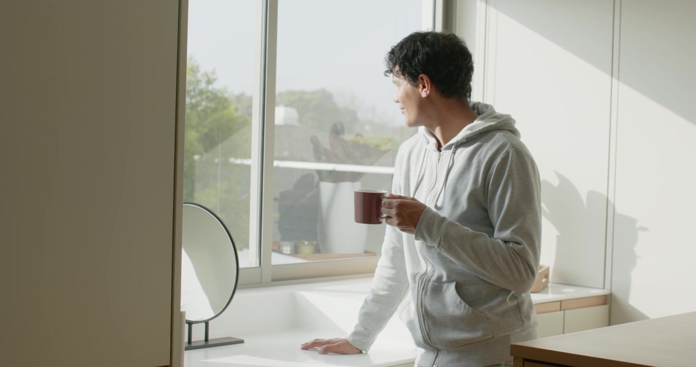 La luce del mattino illumina un uomo a casa, che gira la testa, osserva le piante sul balcone, tenendo una tazza bordeaux
