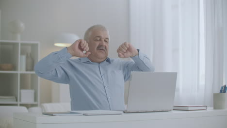 aged man is stretching hands during working with laptop at home feeling overextension and fatigue of muscles feeling bad at workplace