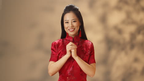 portrait of young asian woman in red traditional clothes smiling cheerfully at camera and doing chinese gesture of congratulation with hands