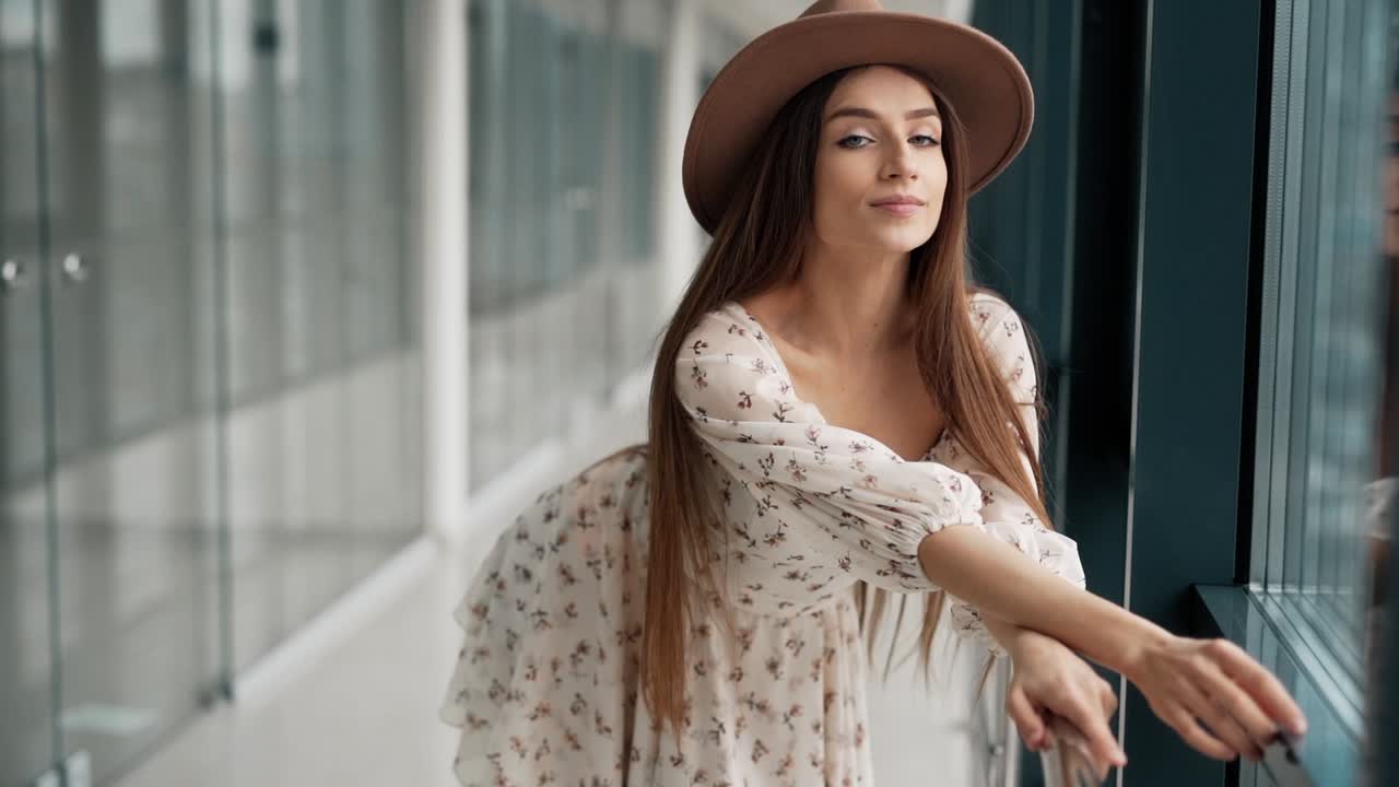 hermosa chica alegre en una tabla y un sombrero se para junto a la ventana en el pasillo y sonríe