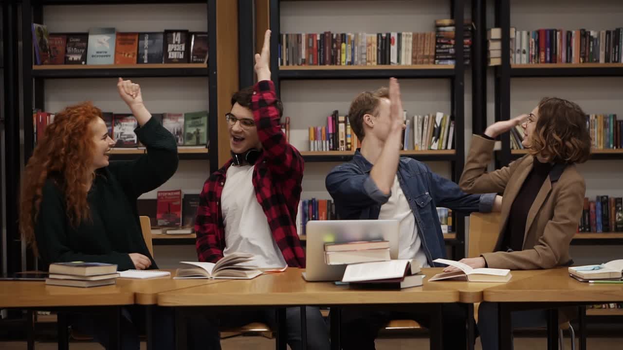 grupo de jóvenes estudiantes hablando usando una computadora portátil explicando el trabajo del curso estudiando juntos en la biblioteca. discutiendo activamente algo, riendo y gesticulando y luego dándose unos a otros cinco. cámara lenta