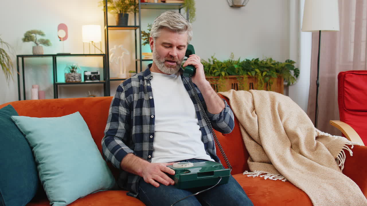 Opprømt, glad moden mann som bruker en gammeldags telefon og nyter samtalen mens han sitter i sofaen hjemme