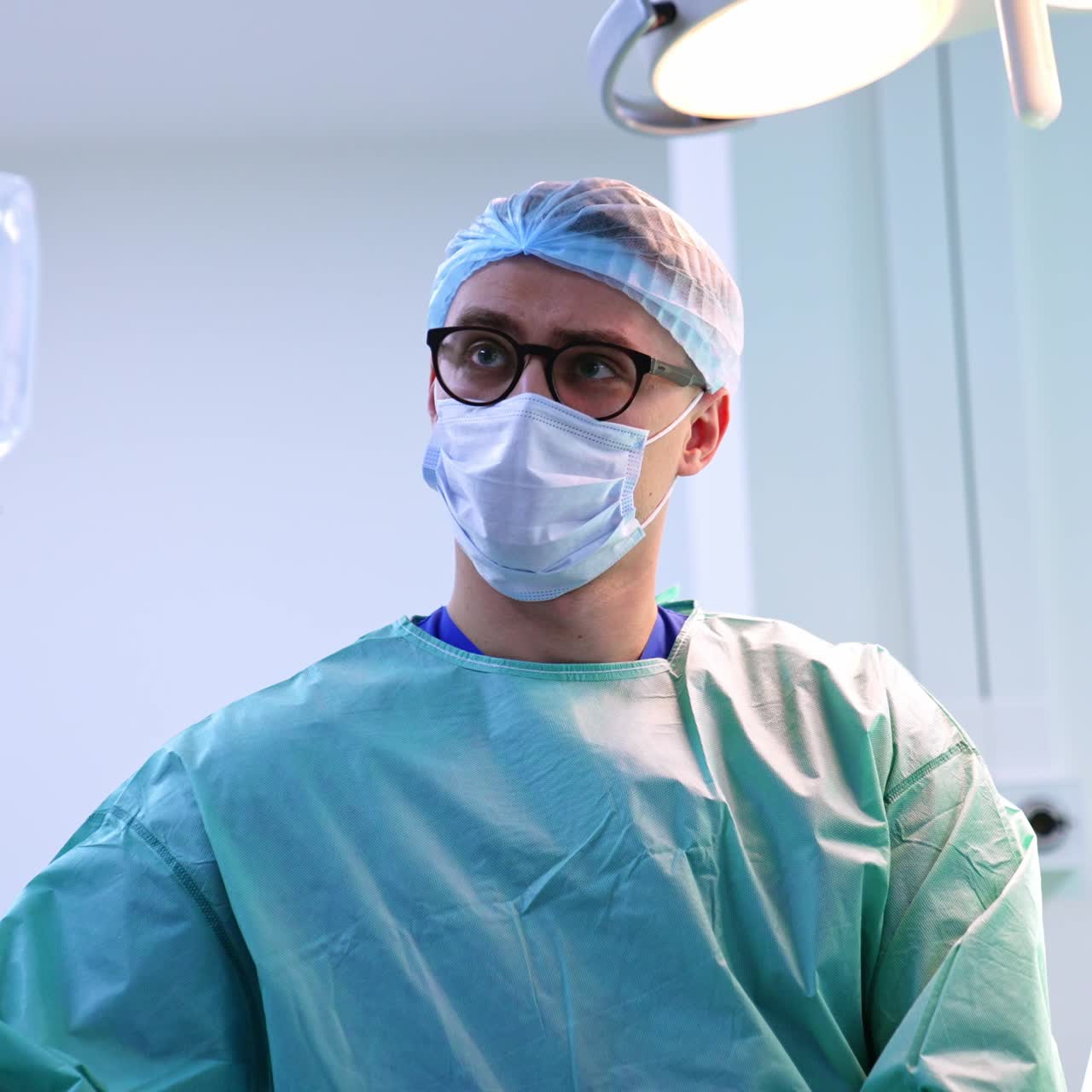 Giovane chirurgo in camice e occhiali in sala operatoria che guarda in alto. Gruppo di medici al lavoro in sala operatoria durante un intervento