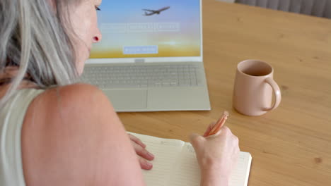 caucasian mature woman with gray hair writing in notebook, looking at laptop