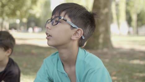 retrato de niño latino con anteojos sentado en la hierba en el parque, escuchando a alguien y hablando