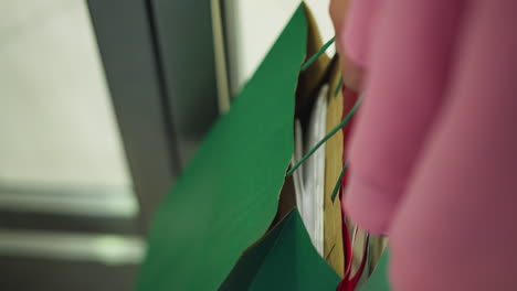 dama llevando bolsas de compras coloridas mientras lleva un vestido rosa, abriendo una puerta para salir de un edificio, una persona con pantalones blancos es parcialmente visible siguiendo detrás, el fondo está borroso