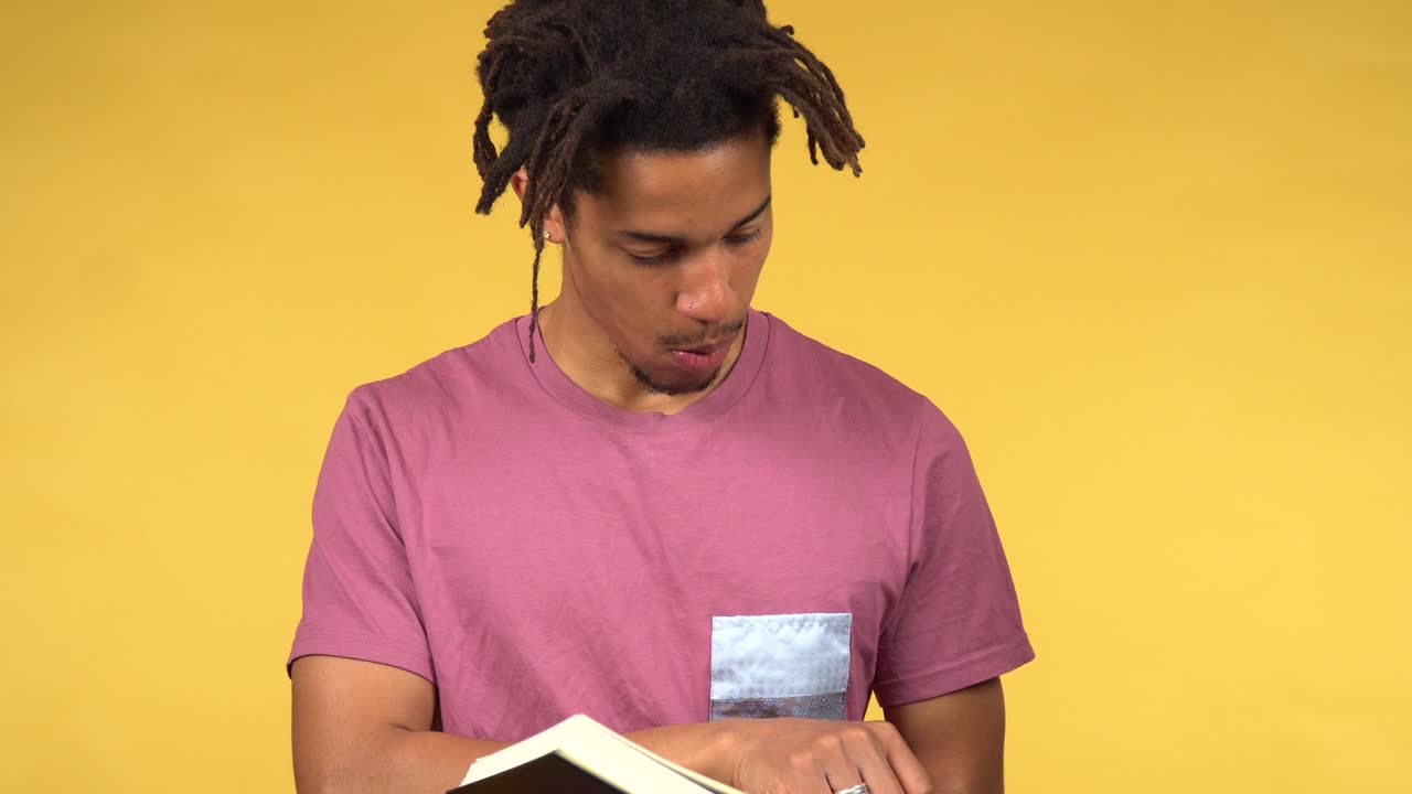 Cheerful young black male student reading a book, yellow background