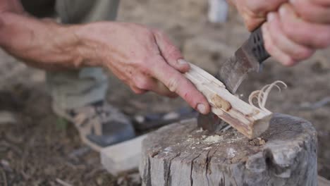 caucasian male survivalist using machete to prepare tinder and kindling wood at camp in wilderness