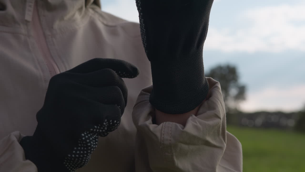 Nærbillede af en person, der justerer en sort handske med et fast greb, ærmerne lynet op, sløret åbent felt og træer i blødt baggrundslys, forbereder sig på action i rolige udendørsomgivelser