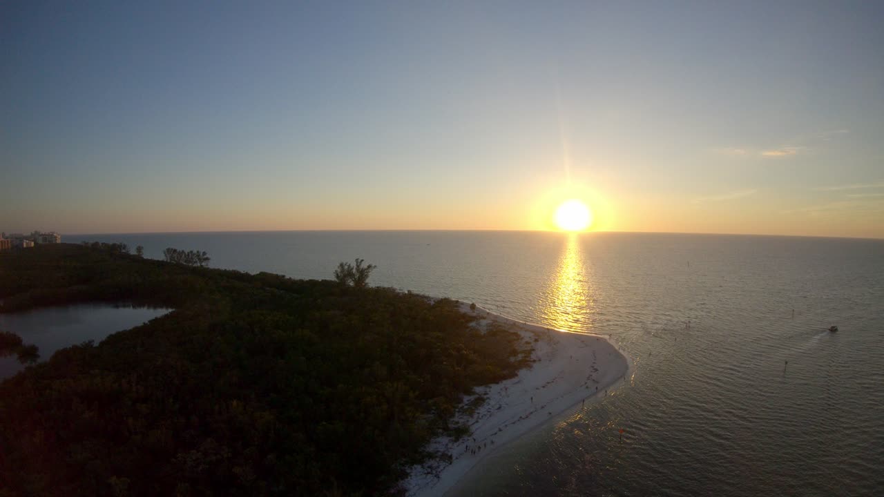 The sun sets over the sea off the coast of Naples. Drone footage moves sideways across the beach and over the inland bodies of water surrounded by palm trees.