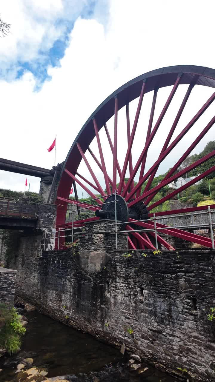 A large red water wheel beside an old stone mill with scaffolding and a bright sky. vertical video