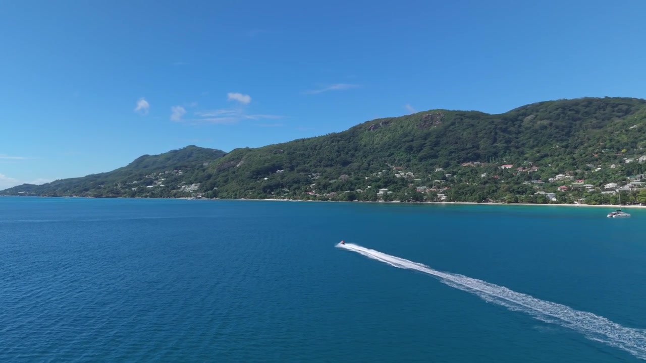 Ampia ripresa aerea con drone di una madre e una figlia su una moto d'acqua che lasciano una lunga e dritta scia bianca attraverso l'oceano blu vibrante di Beau Vallon, Seychelles, con lussureggianti montagne verdi sullo sfondo