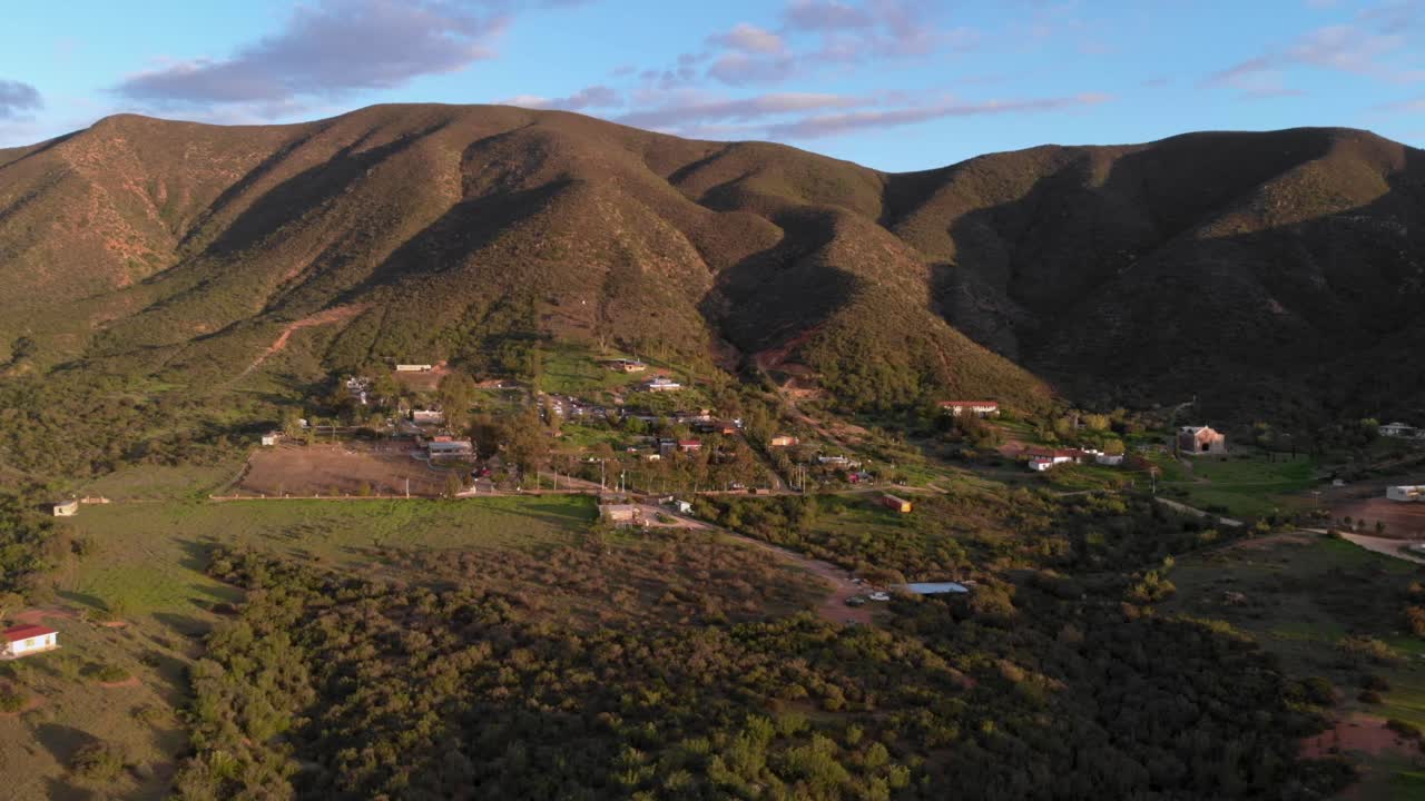 Ripresa aerea con drone di un paesaggio montano con un villaggio collinare, vegetazione verde e case sparse, ampia vista sulla valle che mostra un ambiente rurale e uno scenario naturale.