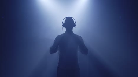 Backlit-silhouette-of-dj-man-standing-in-dark-nightclub-disco,-putting-headphones-on-to-listen-music