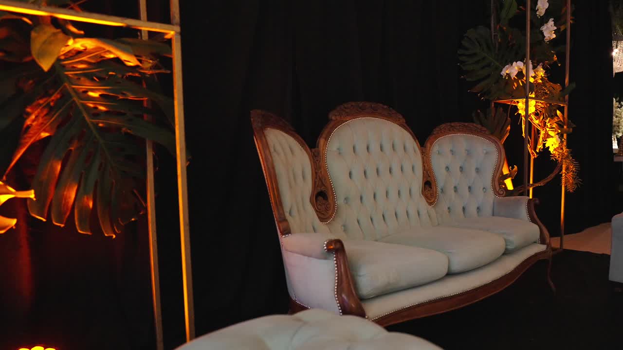 Carrellata in avanti verso un divano in stile vintage su un palco nuziale decorato con composizioni floreali e calda illuminazione ambientale in una location per eventi al chiuso.