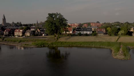 Luftrückwärtsbewegung-Des-Flussufers-Mit-Baum,-Der-Sich-Im-Fluss-Ijssel-Widerspiegelt-Und-Die-Türme-Von-Zutphen-Im-Hintergrund-An-Einem-Bewölkten-Tag-Während-Des-Sonnenuntergangs-Enthüllt