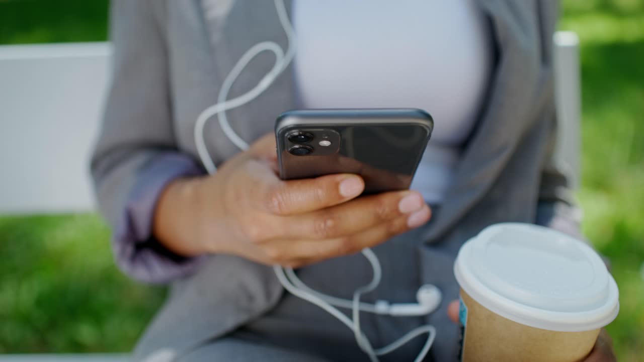 스마트폰을 사용하는 여성이 커피를 마시면서 공원에서 음악을 듣고 있습니다.