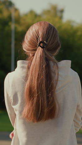 vista trasera de una mujer con una sudadera con capucha y el cabello atado hacia atrás, caminando por una pista vacía del estadio en un día soleado, con los brazos cruzados pensativamente, mientras la cálida luz del sol la ilumina