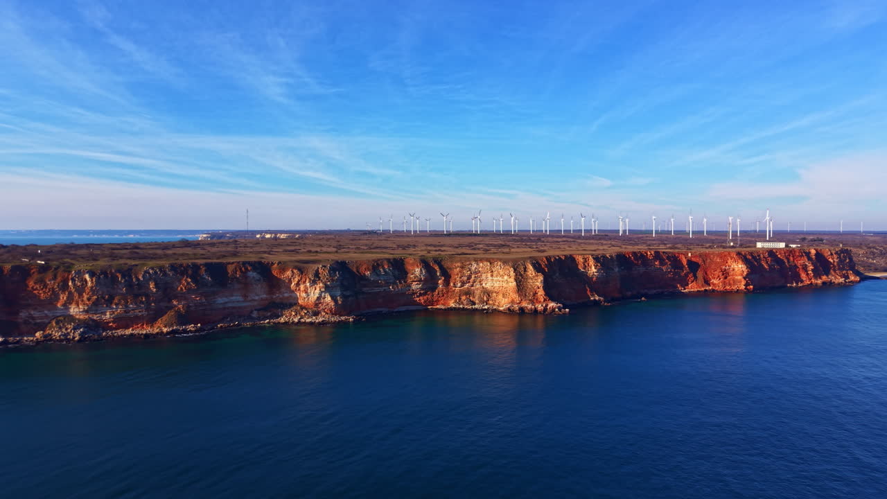 Wind turbines on cliffs above ocean water with blue sky and distant land in view during daytime near coastal area