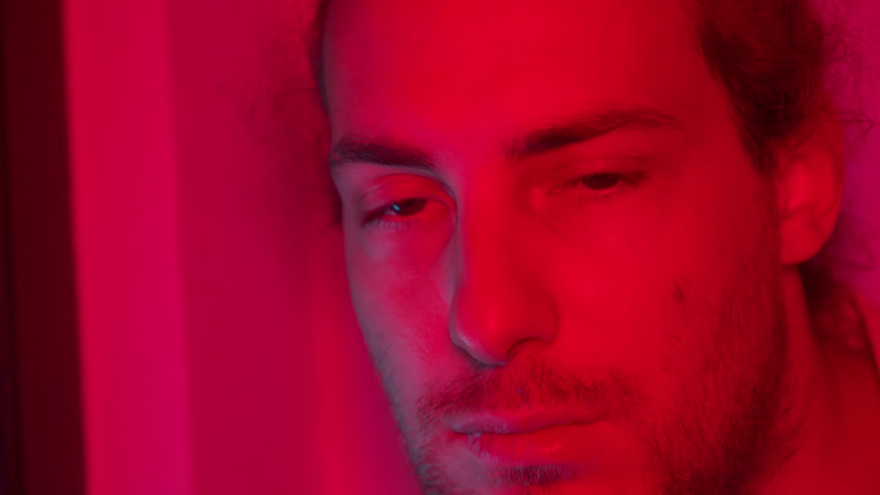 Man under red lighting, calm and serene, eyes closed thoughtfully loooking down and away, peaceful expression, shadows surround