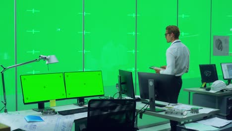 ingeniero masculino guapo de pie en un espacio de oficina con computadoras de escritorio frente a una pared de llaves de pantalla virtual verde con marcadores de lugar para maquetas y plantillas. modelo usando tableta.