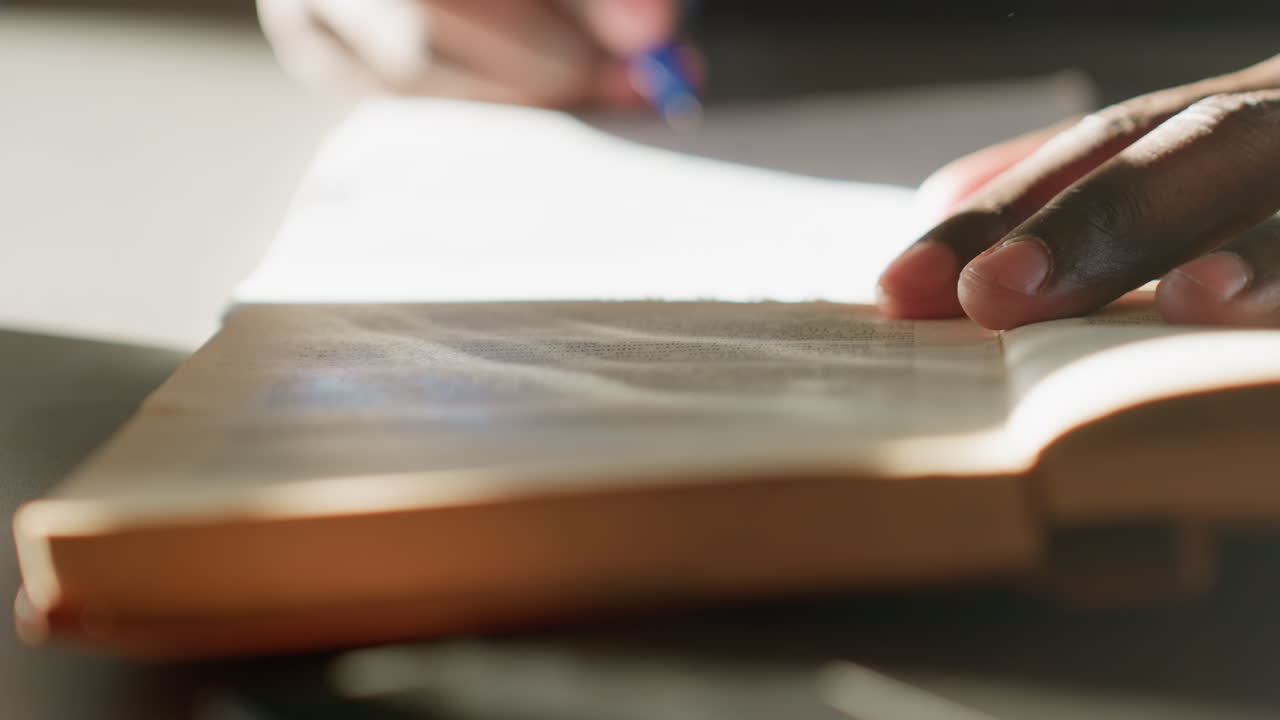 Close up of person flipping through pages of old book with fingers resting on paper, highlighting fine texture, soft warm light, and intimate moment of reading in quiet indoor setting