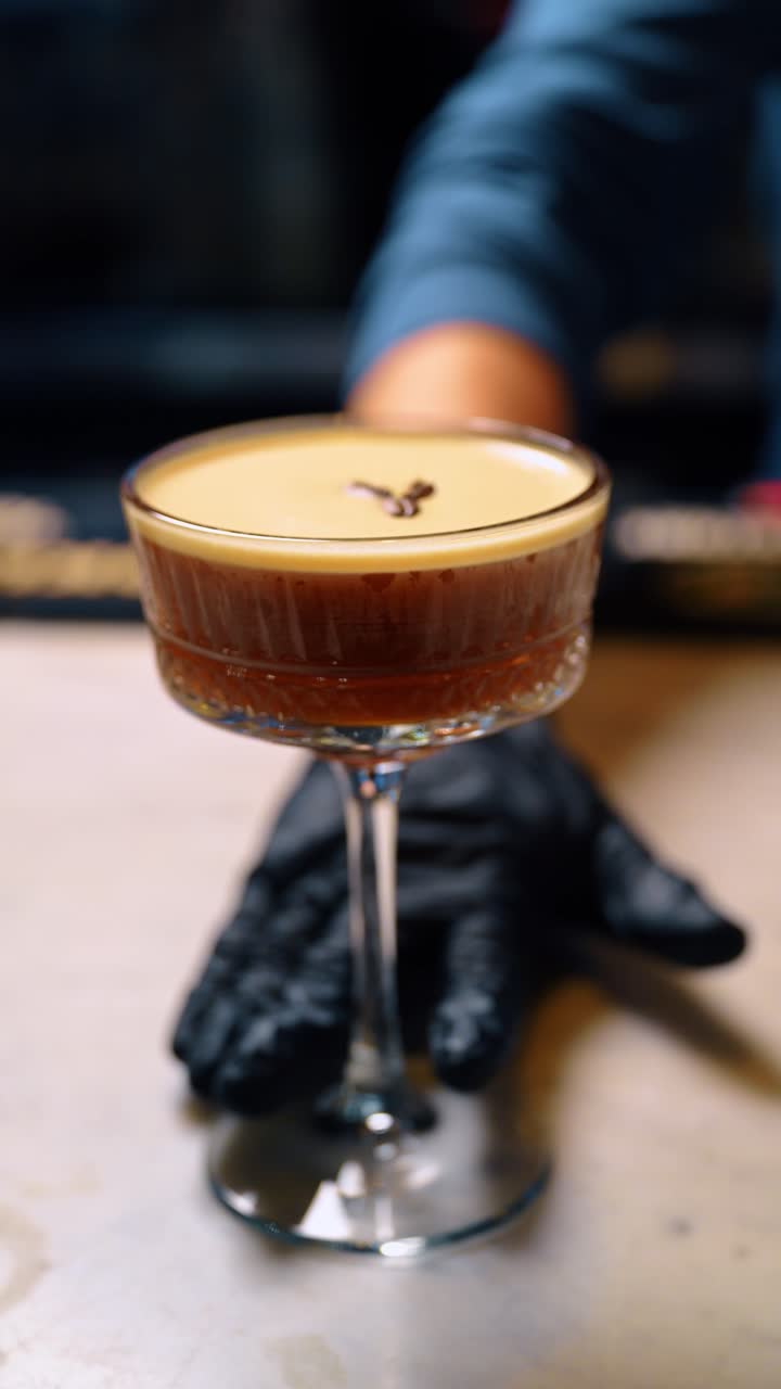 Cocktail Espresso Martini presentato su un bancone da bar in marmo bianco. Bevanda finita con schiuma e chicchi di caffè mostrata con la mano del barista