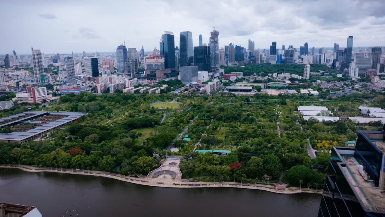 This cinematic drone footage offers a view of a Park in Bangkok, Thailand. The wide expanse of green parkland contrasts beautifully with the surrounding lake and the towering skyline in the distance.