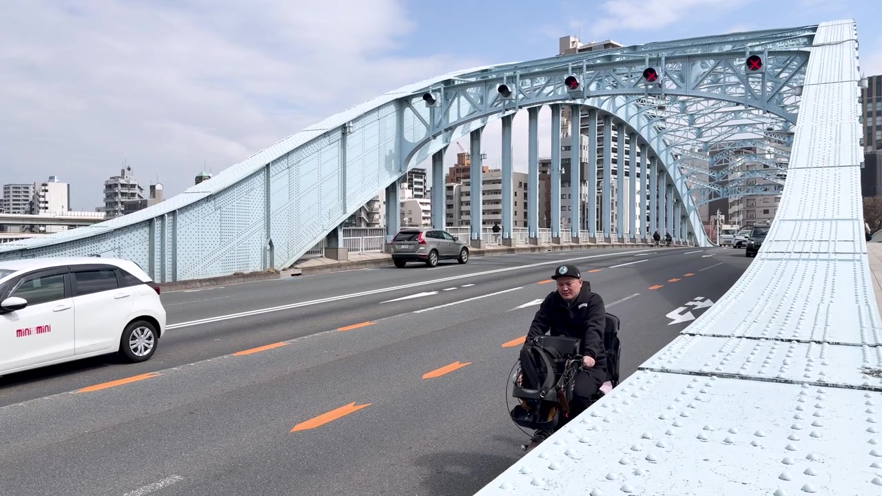 Un uomo su una sedia a rotelle elettrica attraversa un ponte ad arco in acciaio blu a Tokyo