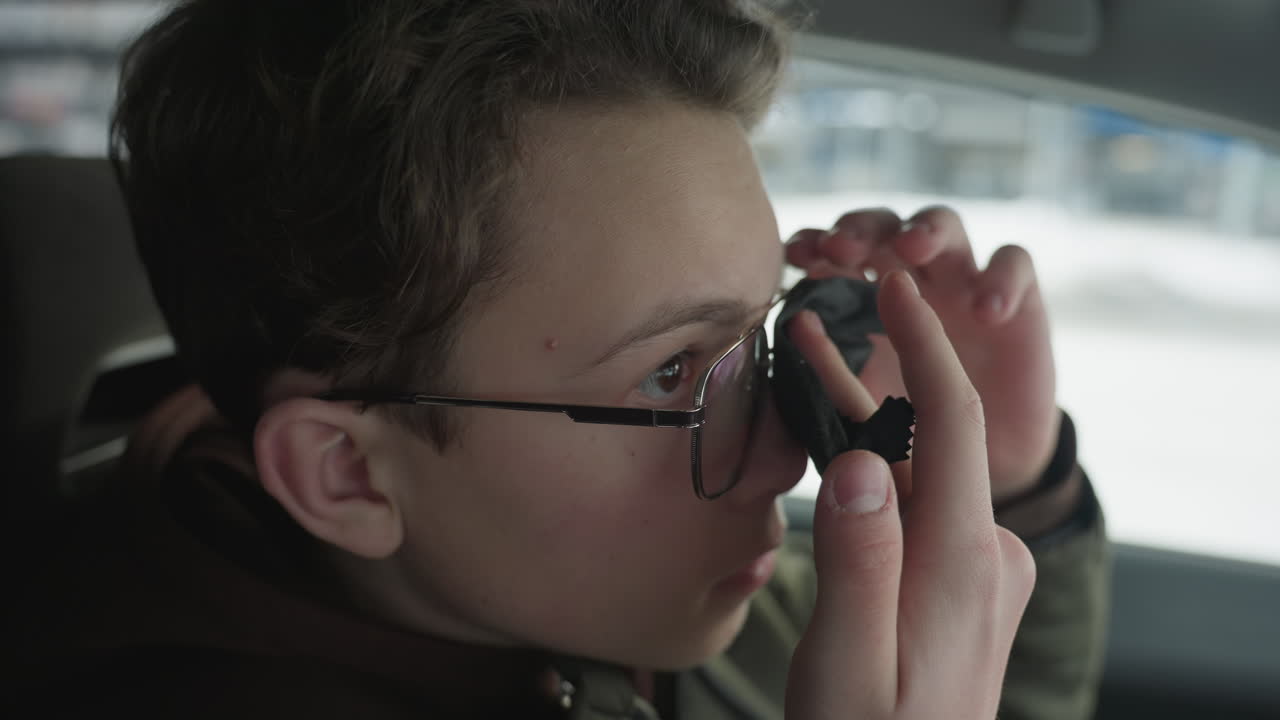 Cute boy cleaning glasses lens with cloth inside car with snowy background blur showing careful hand motion under ambient natural light inside vehicle cabin reflective gaze for eyewear hygiene
