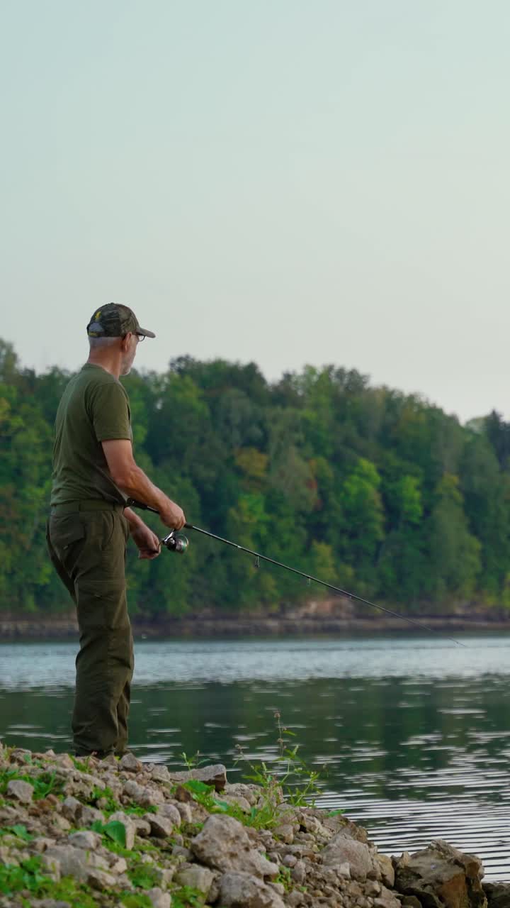 Video vertikal seorang lansia memancing di tepi sungai saat senja. Hutan hijau subur di latar belakang