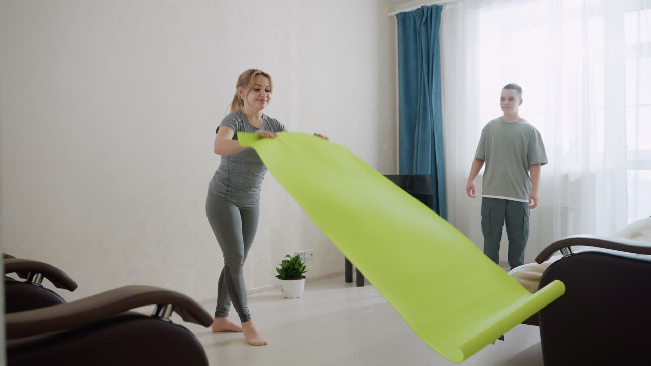 Kid hands over bright green exercise mat to mom in modern living room, both barefoot and dressed in casual sportswear, preparing for workout together with cooperation