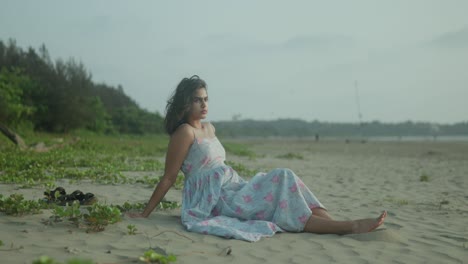 Young-woman-in-a-floral-dress-sitting-thoughtfully-on-a-sandy-beach,-green-foliage-in-background,-overcast-day