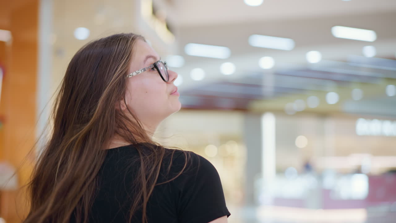 Giovane signora vista da dietro con gli occhiali e lunghi capelli che le ricadono sulle spalle, sorridente e che guarda di lato in un centro commerciale ben illuminato, che trasuda sicurezza e felicità