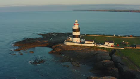 Aerial-view,-sunrise,-low-pan-left,-Hook-Lighthouse-is-situated-on-Hook-Head-at-the-tip-of-the-Hook-Peninsula-in-Co-Wexford,Ireland,-oldest-lighthouse-in-the-world,-was-built-in-the-12th-century