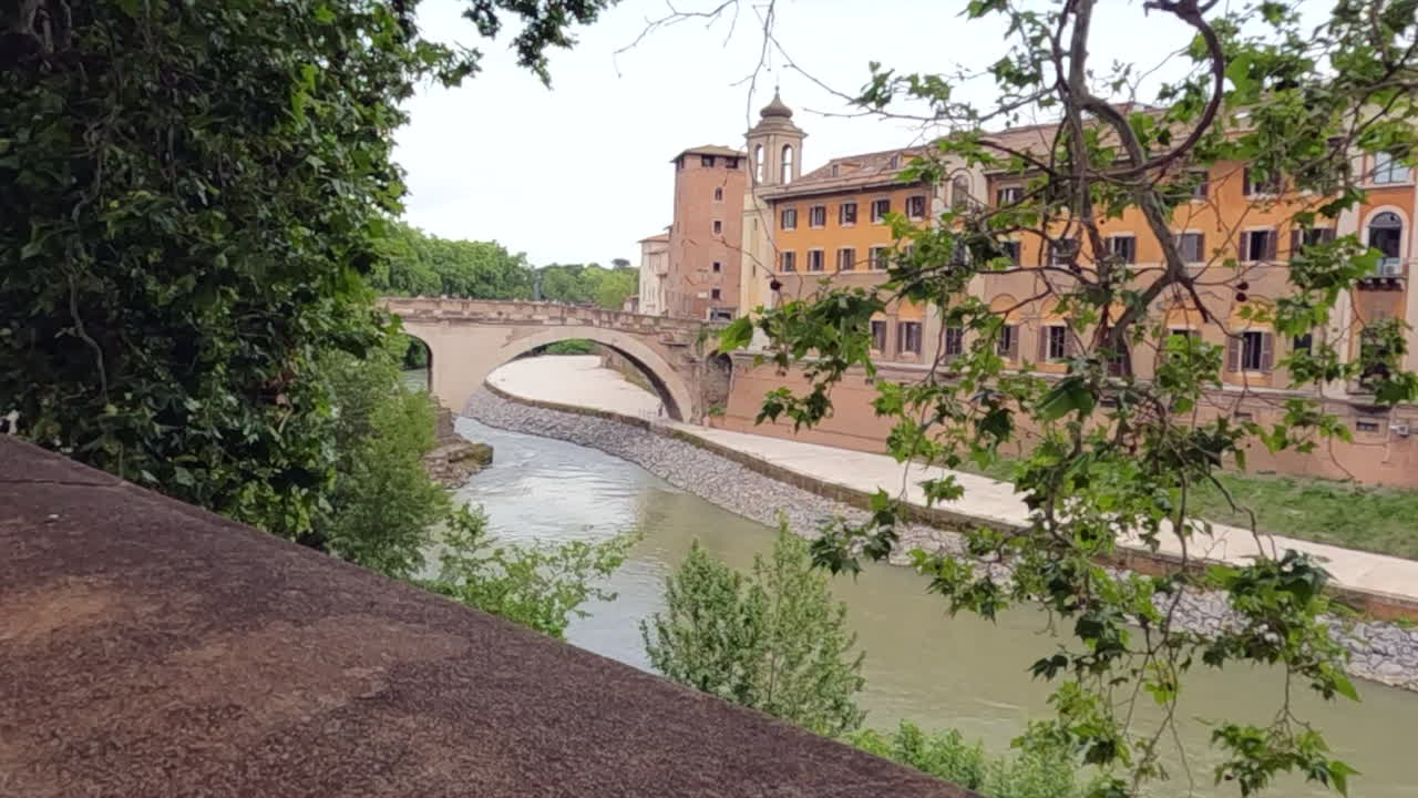 Tiber Nehri'ni geçen antik Roma köprüsünün, tarihi toprak renkli binalar ve gür yeşil ağaçlarla nefes kesen manzarası. Roma'nın ebedi şehrinde ikonik İtalyan mimarisi ve doğal sahil şeridi.