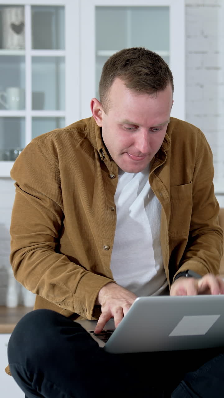 Funny guy sitting on a kitchen table and typing on a laptop. Young fellow studying on a laptop for long period. Tired face of a freelancer working at home. Remote job. Vertical video