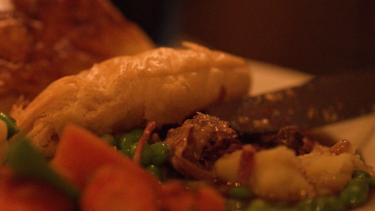 Steak pie being sliced, served with peas, carrots, and mashed potatoes under warm lighting