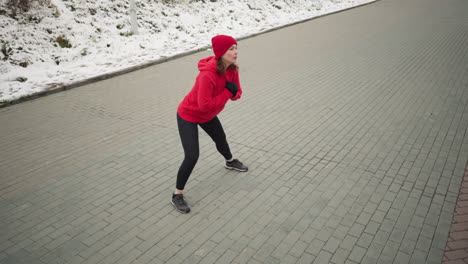 woman performing outdoor winter workout, stretching into a deep side lunge on paved ground, arms clamped close to chest, dressed in red hoodie and black leggings, near snowy hill in cold environment