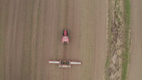 overhead, look down view from drone tracking large red farm tractor towing machinery as farmer works in field to prepare land for planting