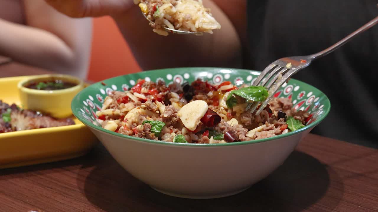 Person uses spoon and fork to eat spicy beef, garlic, and chili rice in a casual Bangkok restaurant. Warm lighting, close-up, steady camera
