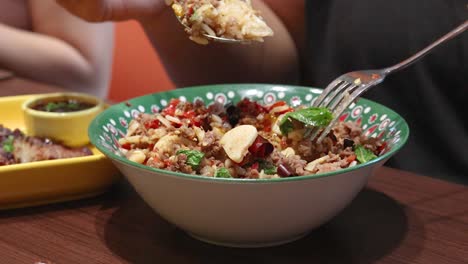person uses spoon and fork to eat spicy beef, garlic, and chili rice in a casual bangkok restaurant. warm lighting, close-up, steady camera