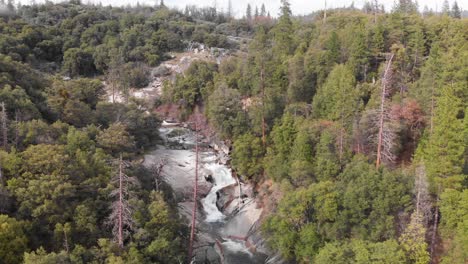 Toma-Aérea-Giratoria-De-Una-Cascada-En-Un-Escarpado-Bosque-De-Pinos-Y-Robles-En-California