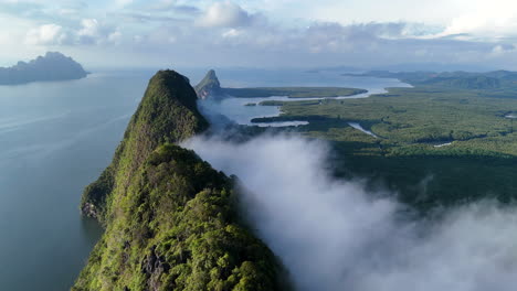 dramatic cliffs rise as mist floats into air from forested ridge jungle in phuket, thailand during golden hour, aerial fly through