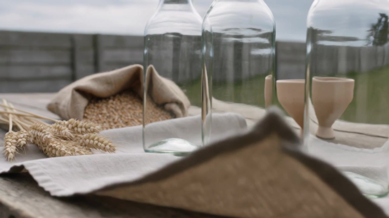 Shifting camera approaching three glass bottles on rustic table, showing burlap sack, wheat, linen