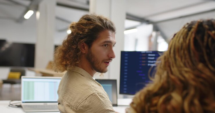 Diverse coworkers watching male dev reviewing code and checking monitors and laptop at office desk